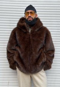 Cappotto di pelliccia marrone con cappuccio, dotato di chiusura a zip e tessuto spesso. Abbinato a pantaloni beige con motivo a fantasia.