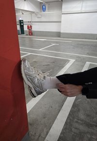 Λευκό αθλητικό παπούτσι New Balance με υφή ασημί πάνω μέρος, δεμένο με λευκές κορδόνες, τοποθετημένο σε έναν κόκκινο τοίχο σε γκαράζ.