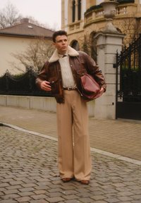 Veste en cuir marron avec col en peau lainée, pull en tricot torsadé beige, pantalon beige à jambes larges, chaussures marron, tenant un sac bordeaux.