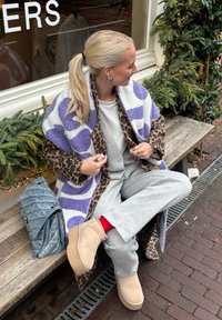 Casaco oversized com padrão de estampa de onça e bolinhas roxas, calças de moletom cinzentas, blusa creme, botas bege, e uma mala azul acolchoada.