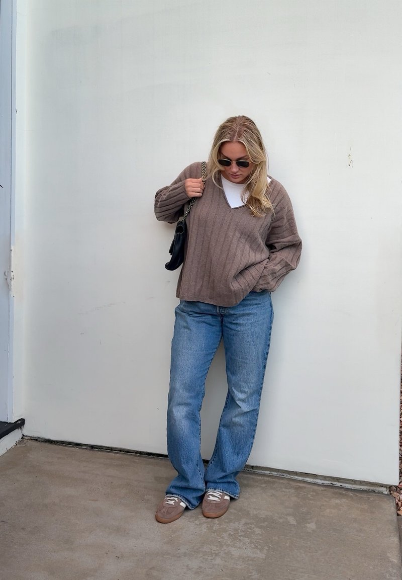Mujer con gafas de sol, suéter marrón, vaqueros azules y zapatos marrones, de pie contra una pared blanca sosteniendo un bolso negro de hombro.