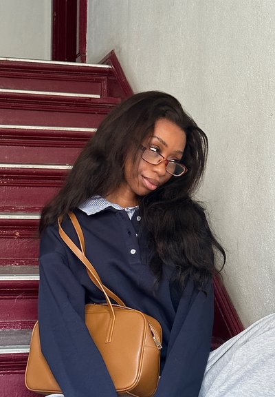 El modelo lleva una camiseta polo de manga larga en color marino con un cuello a rayas y un bolso bandolera marrón. Al fondo se ven escaleras rojas y una pared lisa.