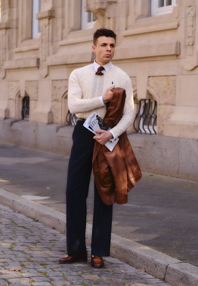 Un hombre con un suéter de punto en color crema sostiene una chaqueta de cuero marrón y una revista. Lleva pantalones azul marino y zapatos marrones, de pie sobre adoquines.