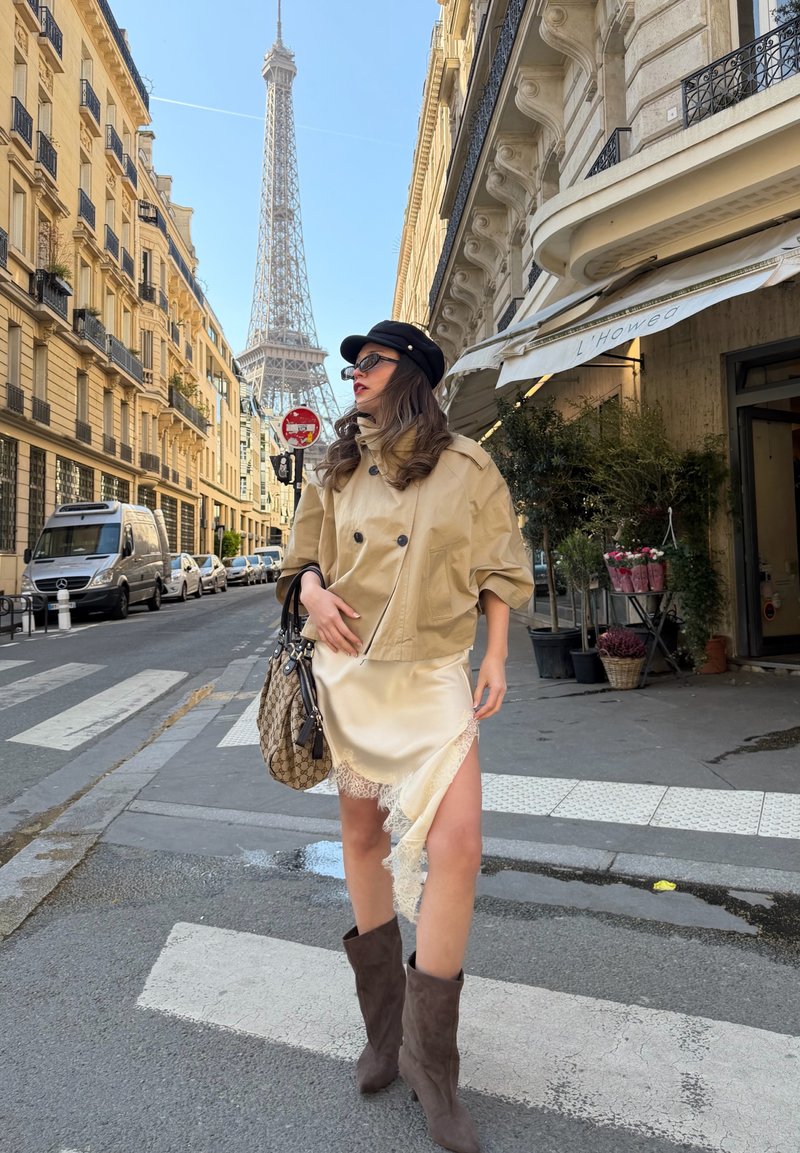 Femme portant une veste beige, une robe en dentelle crème et des bottes marron, debout dans une rue de Paris avec la Tour Eiffel en arrière-plan.