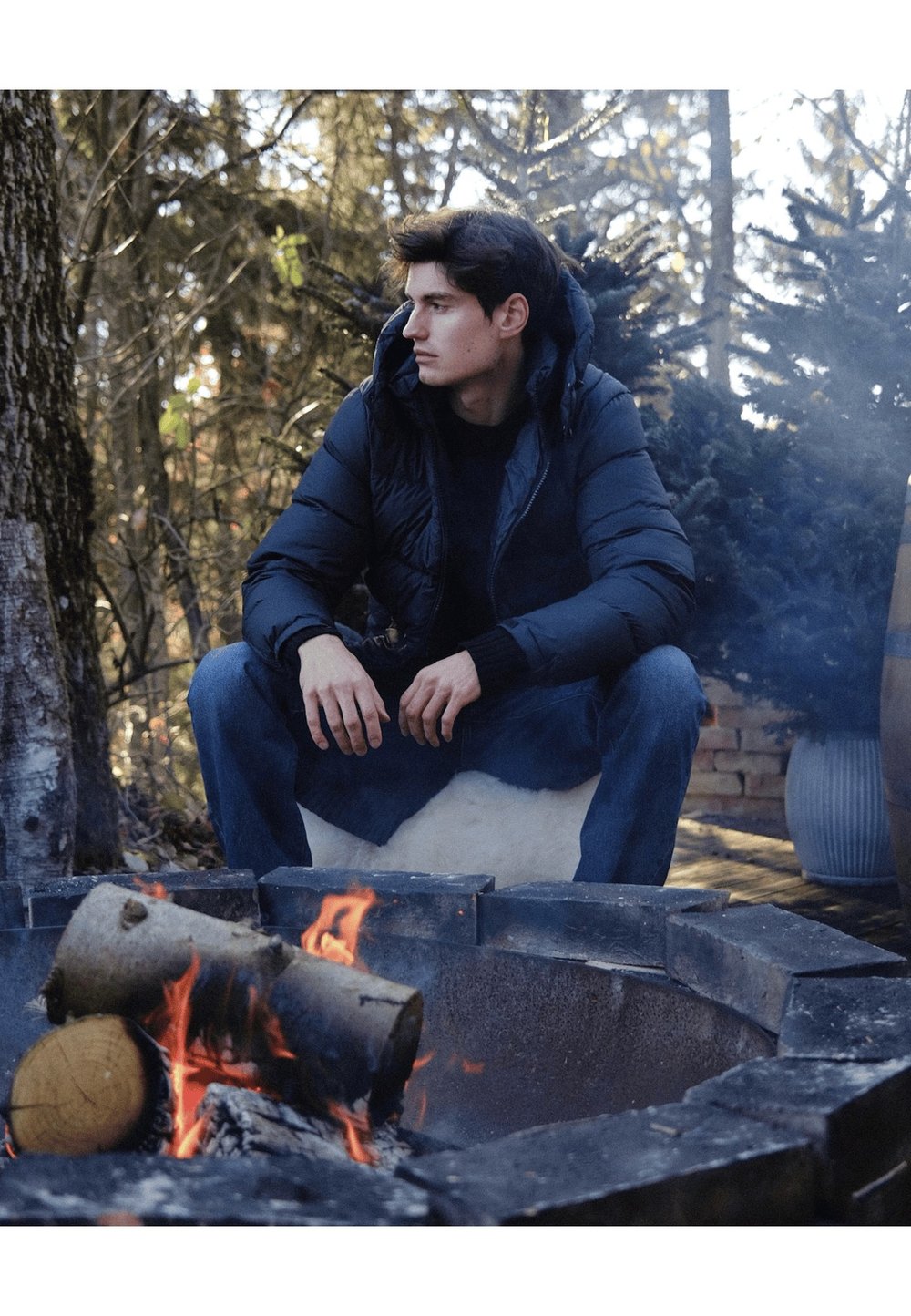 Melnā pūderjakā ar kapuci, vilkta virs tumša krekla. Ainā redzama ugunskura vieta ar baļķiem un dabīga koku aizmugure.