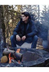 Melnā pūderjakā ar kapuci, vilkta virs tumša krekla. Ainā redzama ugunskura vieta ar baļķiem un dabīga koku aizmugure.