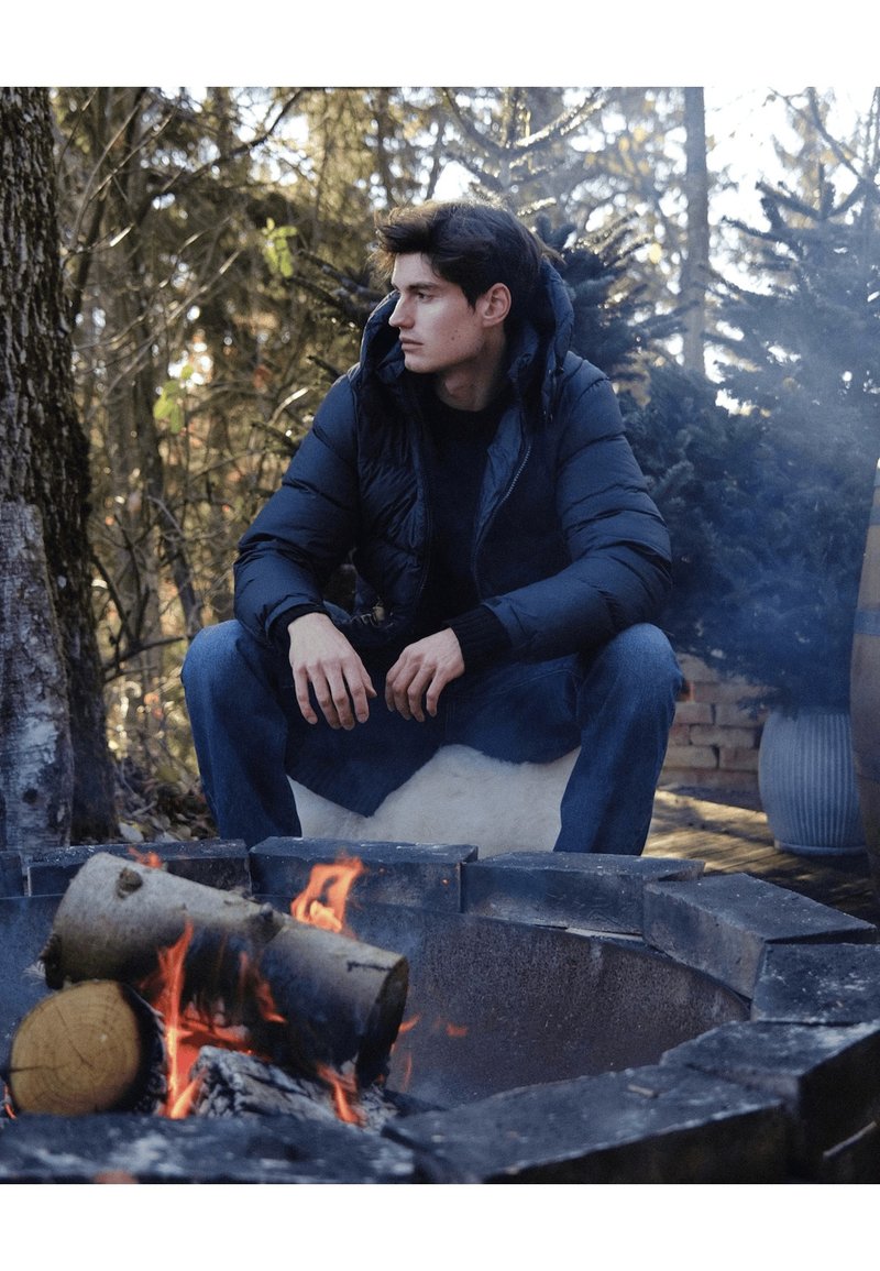Doudoune noire à capuche portée sur une chemise foncée. La scène comprend un foyer avec des bûches et un arrière-plan naturel d'arbres.