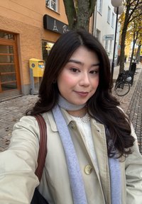 Nuori tummahiuksinen nainen beigen värisessä takissa ja harmaassa huivissa ottaa selfien mukulakivikadulla, jonka varrella on syksyisiä puita ja polkupyöriä.