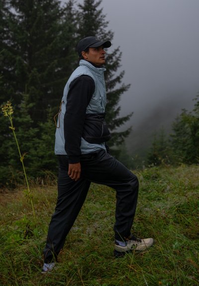 Chaleco sin mangas azul claro sobre una camisa negra de manga larga, pantalones negros y zapatos beige; de pie en la hierba con árboles en un fondo neblinoso.