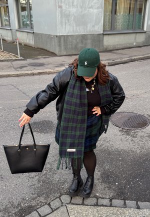 Schwarze Lederhandtasche, grüner Hut, kariertes Tuch in dunklen Blau- und Grüntönen, schwarze Stiefeletten und ein schwarzes Outfit. Gehweg-Setting.