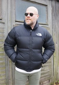 Zwart pufferjack met een hoge kraag, ritssluiting aan de voorkant en wit logo op de linkerborst. Getextureerde, gestikte stof met elastische manchetten.