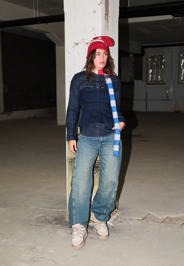 Une femme porte un bonnet rouge en maille, une écharpe rayée, une veste en jean, un jean large et des baskets blanches dans un intérieur industriel peu éclairé.