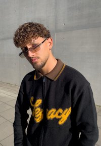 Črni zip-up pulover z rumenim napisom "Legacy", rjavim ovratnikom in teksturirano tkanino. Model nosi sončna očala s filtrom. Betonska podlaga.