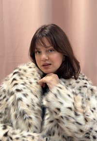 Femme aux cheveux bruns mi-longs portant un manteau en fausse fourrure à pois, touchant légèrement le col, sur un fond rose pâle.