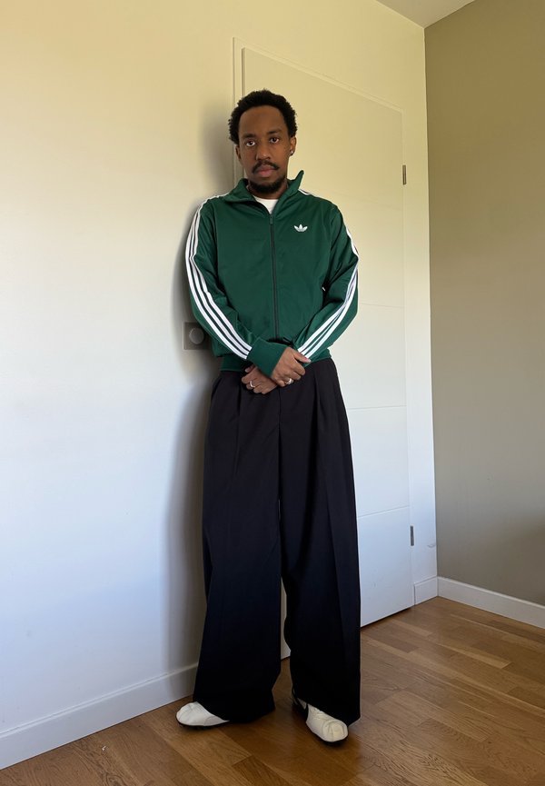 Un hombre está de pie en un interior con las manos juntas. Lleva una chaqueta deportiva verde de rayas, unos pantalones negros anchos y zapatos blancos.