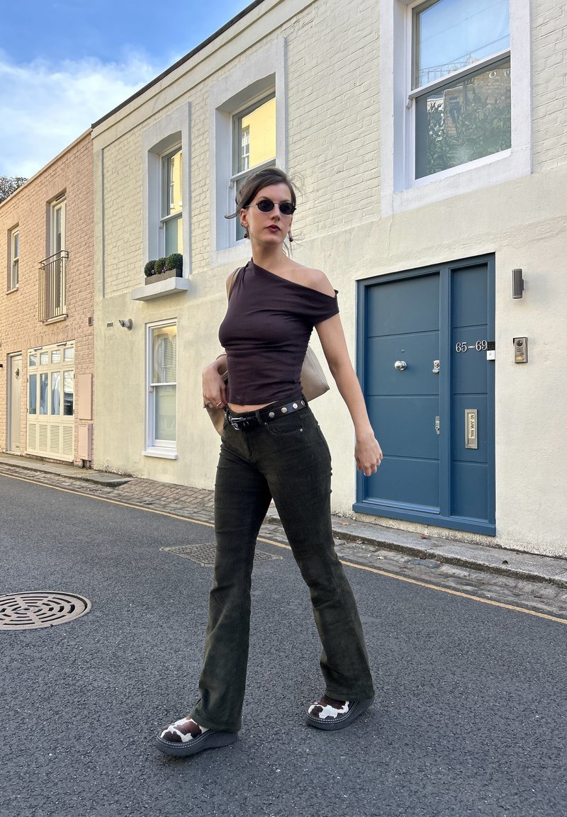 Femme marchant dans la rue, portant un top noir à une épaule, un pantalon sombre, des chaussures à motif vache, de petites lunettes de soleil noires et portant un sac beige sur l'épaule.
