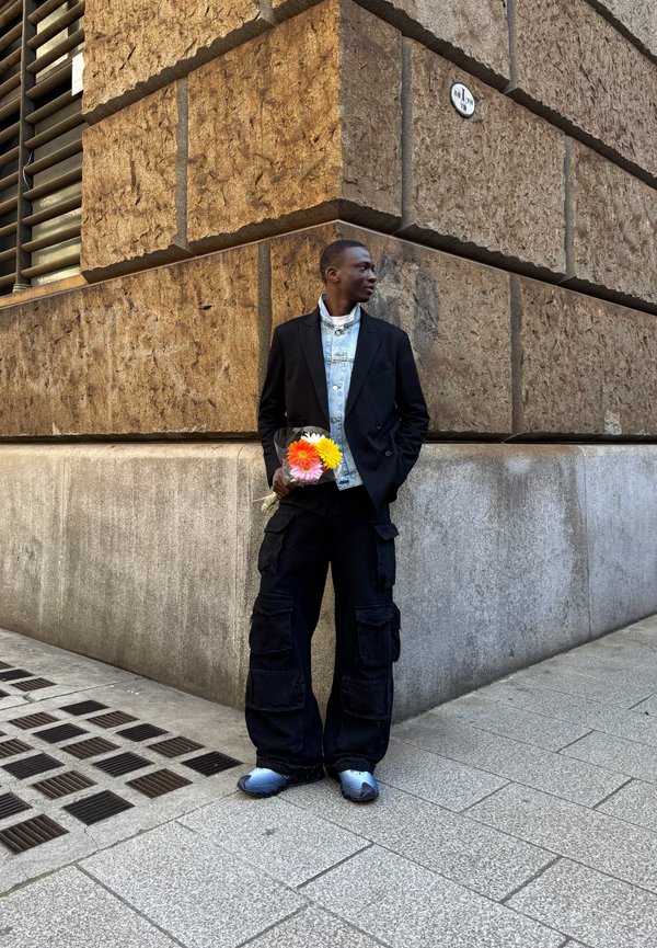 Schwarze Blazer über einer hellen Jeansjacke, schwarze Cargo-Hose und graue Sneaker, die einen Strauß bunten Blumen vor einer strukturierten Wand halten.