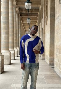 Pull en maille bleu et crème, avec un motif ondulé, accompagné d'un jean délavé clair à texture diamantée. En intérieur, devant une colonne en pierre.