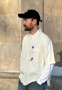 Homme barbu portant une casquette noire, une chemise crème à manches courtes par-dessus un sous-pull blanc à manches longues, un jean bleu, debout contre un mur en pierre, les mains dans les poches.