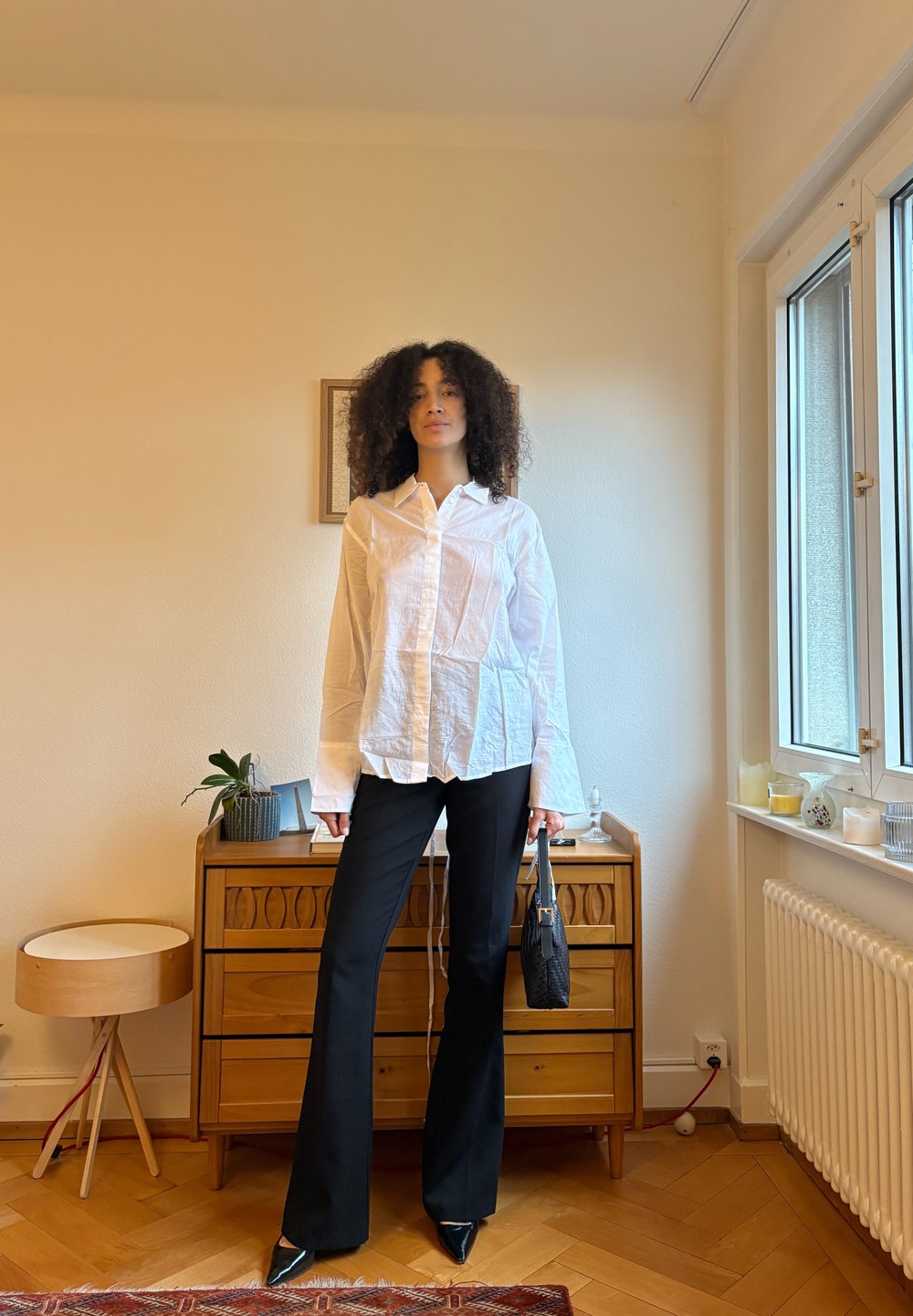 White long-sleeve button-up shirt with textured fabric, paired with black flared trousers and pointed black shoes, holding a small black handbag.