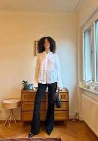 White long-sleeve button-up shirt with textured fabric, paired with black flared trousers and pointed black shoes, holding a small black handbag.