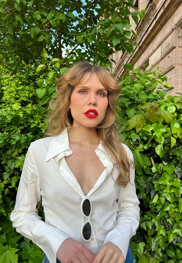 Chemise blanche à boutons avec un décolleté en V profond, associée à des lunettes de soleil noires. Des cheveux blonds ondulés encadrent le visage, sur fond de feuillages verdoyants.