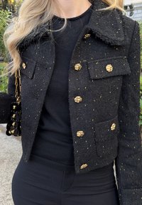 Giacca in tweed nera con bottoni dorati e sottili accenti gialli, dotata di colletto e tasche a toppa, abbinata a un top nero.