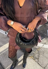 Personne en robe violette transparente et collants imprimé léopard tenant un sac à main noir matelassé, portant plusieurs bagues, une montre en or et des bottes noires.