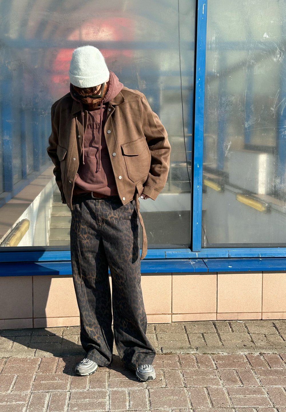 Brown jacket over a maroon hoodie, patterned black trousers, grey trainers, and a light grey beanie; standing outdoors near a blue-framed shelter.
