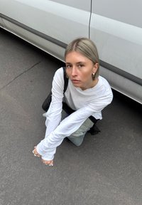 Joven con cabello rubio y camiseta blanca de manga larga agachada en la acera cerca de un vehículo blanco, mirando hacia la cámara.