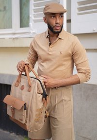 Beige batoh s hnědými koženými akcenty, hlavní přihrádka na zip, přední klopa a boční popruhy. Model má na sobě odpovídající béžové polo tričko a čepici.