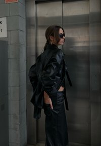 Μαύρο μπουφάν από τεχνητό δέρμα με υπερμεγέθη σχέδιο και μπροστινές τσέπες, συνδυασμένο με ψηλοκάβαλο μαύρο δερμάτινο φούστα και γυαλιά ηλίου.