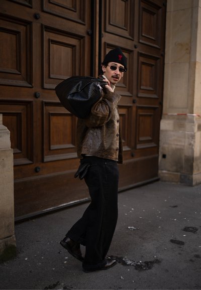 Hombre con gafas de sol y gorro, vestido con una chaqueta de cuero marrón, llevando una gran bolsa negra sobre el hombro, caminando junto a unas puertas de madera.
