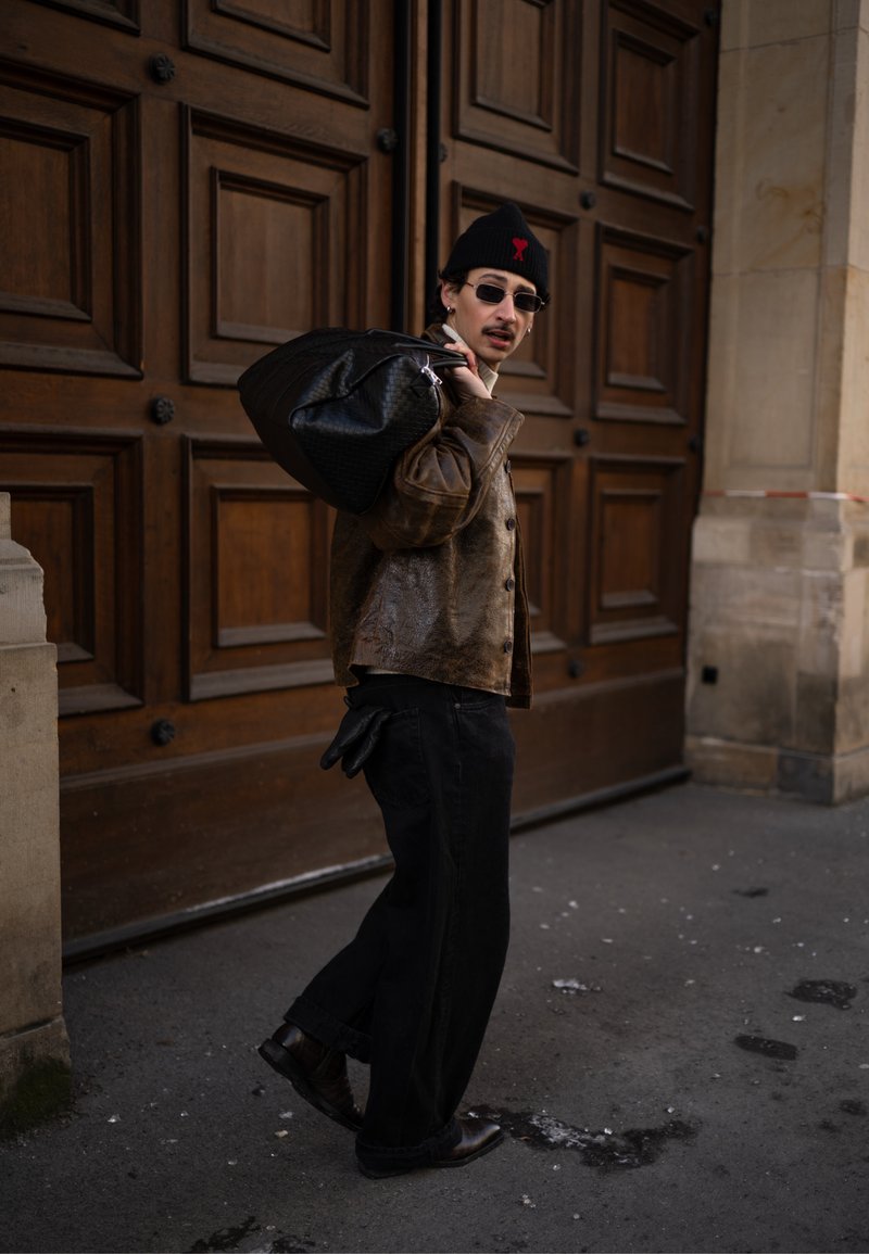 Muž v slnečných okuliaroch a čiapke s hnedou koženou bundou, nesúci veľkú čiernu tašku cez rameno, kráča okolo drevených dverí.