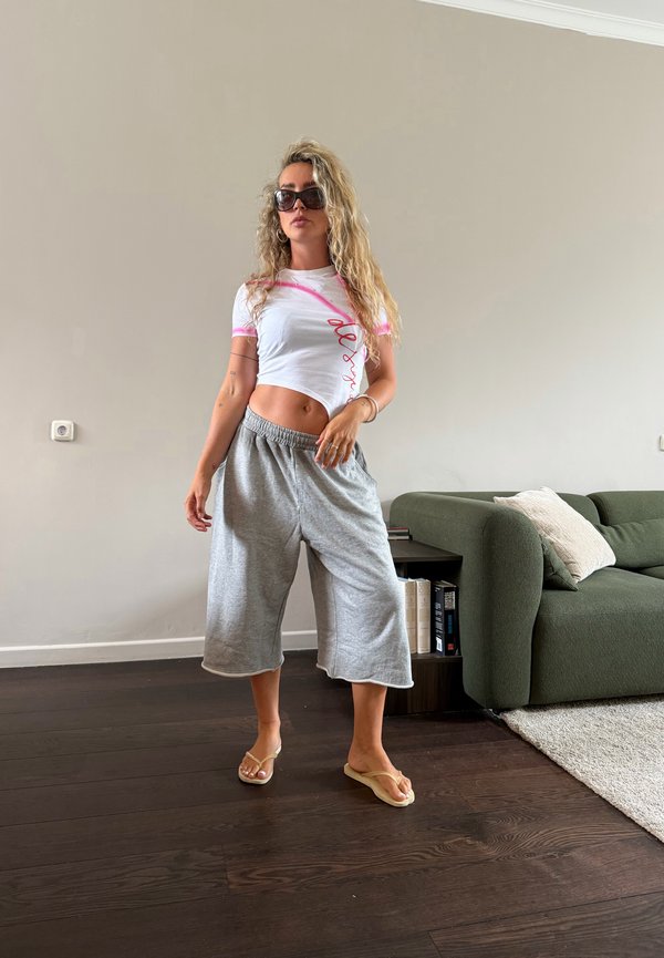Jeune femme aux cheveux bouclés portant des lunettes de soleil, un crop top blanc, un pantalon large gris et des tongs, debout dans un salon avec un canapé et des livres.