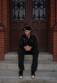 Leren jas, zwart shirt, zwarte spijkerbroek en zwarte sneakers met witte accenten. Zittend op stenen traptreden met een sierlijke houten deur op de achtergrond.