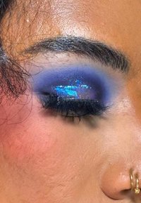 Nærbilde av et øye med mørk blå og lilla øyeskygge, prydet med glitrende accenter. Inkluderer markante vipper og en gullring i nesen.