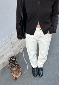 Sort strikket cardigan med knapper, cremefarvede vidde bukser, sorte loafers og en lille hund i en leopardmønstret sweater i snor.