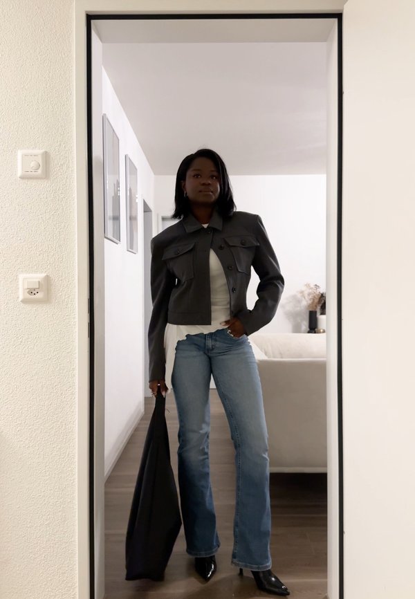 Frau in einer dunklen, kurzen Jacke, weissem Hemd, blauen Jeans und schwarzen Absätzen steht in einem Türrahmen und hält eine schwarze Tasche in einem modernen Raum.