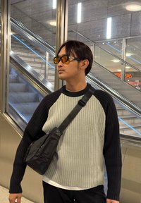 Pull gris et noir à côtes avec manches longues, porté sur une chemise blanche. Un sac bandoulière noir avec des fermetures éclair est posé sur l'épaule.