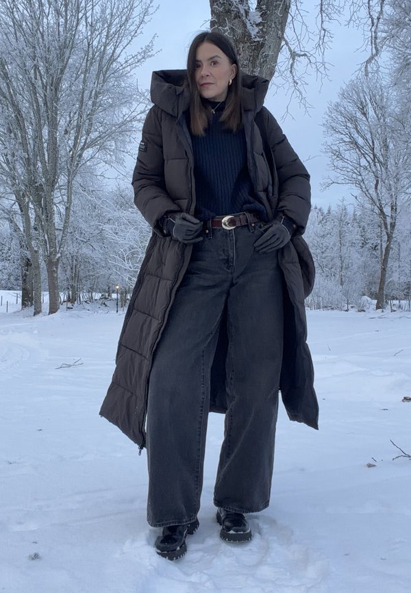 Femme portant un long manteau sombre, un large jean noir, des gants et des bottes, debout dans un paysage enneigé avec des arbres couverts de gel.