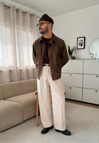 Veste marron à boutons portée sur un pull foncé, associée à un pantalon large couleur crème et des chaussures noires. Cadre intérieur avec meubles visibles.