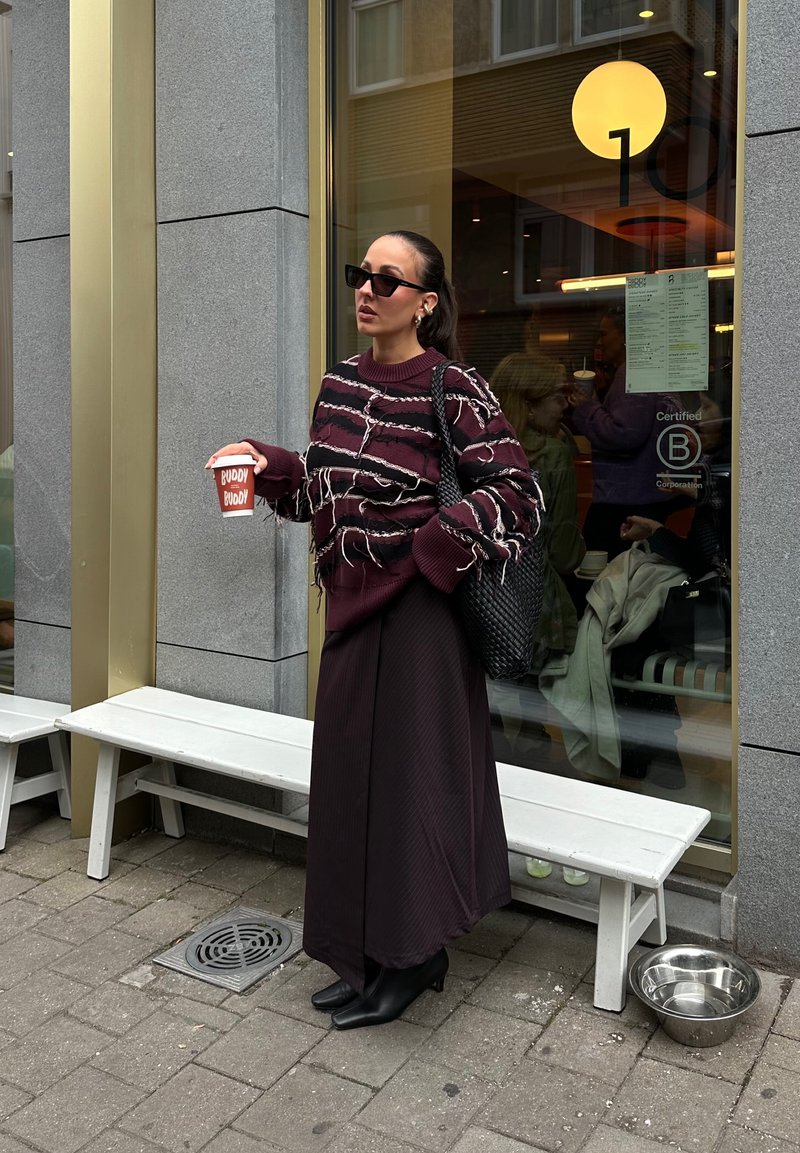 Kvinde i en revnet stribet sweater og lang nederdel, der holder en kaffekop, stående udenfor en café med en bænk og en vandskål til kæledyr på fortovet.
