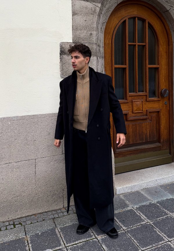 Un homme en long manteau noir et en pull beige se tient à côté d’une porte en bois cintrée, dans une rue pavée.