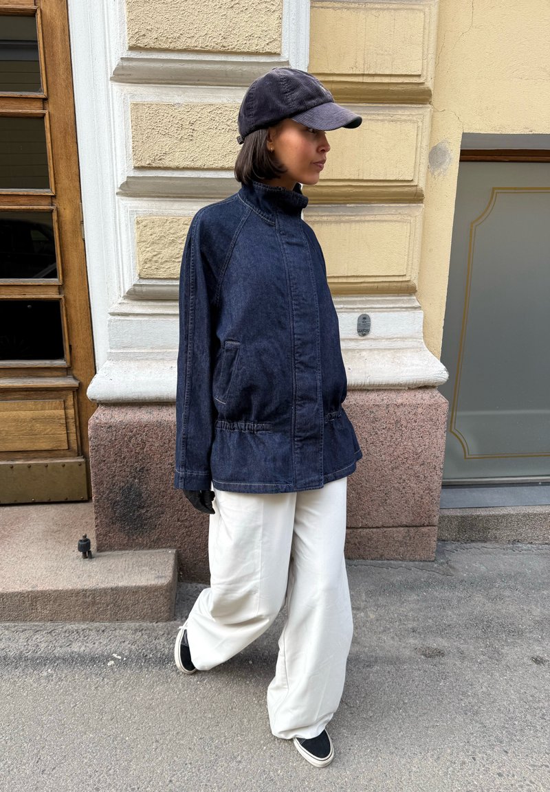 Personne marchant en plein air portant une veste en jean foncé, une casquette noire, des gants noirs, un pantalon blanc ample et des chaussures noires sans lacets.