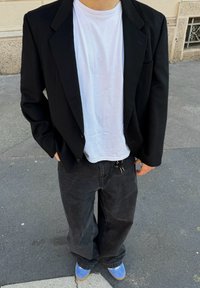 Blazer preto sobre uma t-shirt branca, combinado com calças de ganga pretas e sapatos azuis. O conjunto apresenta um corte descontraído e um design simples.