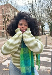 Πράσινη και άσπρη ριγέ ζακέτα με χαλαρή εφαρμογή και oversized μανίκια, συνδυασμένη με ένα κασκόλ σε καρό σχέδιο μπλέ και πράσινου. Αστική υπαίθρια τοποθεσία.