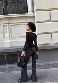 Schwarzes, durchsichtiges Langarmtop mit gerafftem Rücken, kombiniert mit dunklen, ausgestellten Jeans. Die Accessoires umfassen eine mit Nieten besetzte schwarze Handtasche und eine Sonnenbrille.