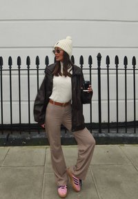 Casaco oversized castanho escuro, suéter branco com textura em nervuras, calças bege com corte flared, sapatos rosa com riscas azuis. Modelo segurando uma chávena de café, usando um gorro branco e óculos de sol.