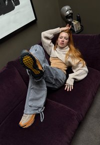 Beige knitted sweater layered over a lighter top, loose-fitting grey cargo trousers, and tan sneakers. Accessories include several rings. Dark purple sofa in the background.